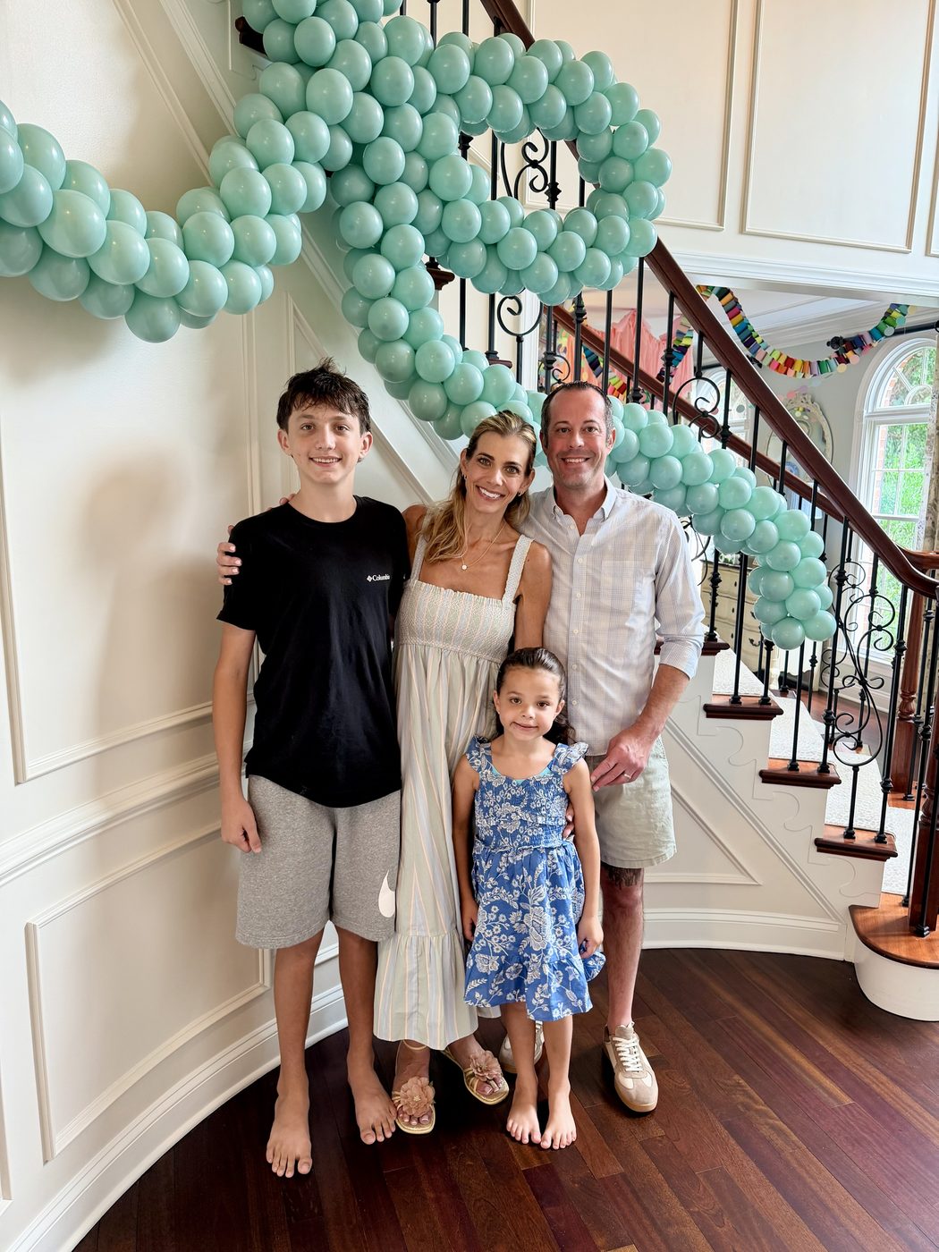 Natalie Wildemore with her family at home, standing beneath a sage-green balloon garland that winds up the staircase