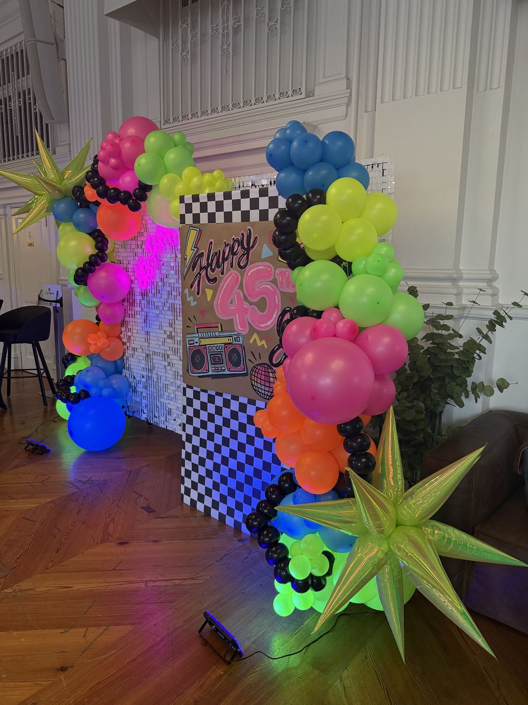 Neon balloon garland around a shimmer wall and checkered panel for a 45th birthday under glowing lights