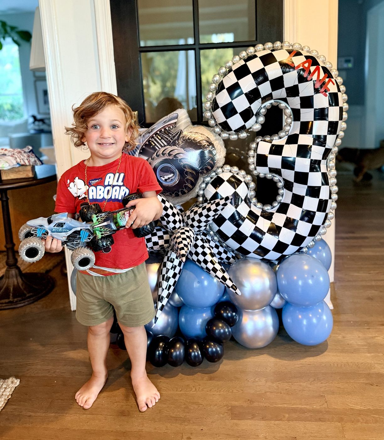 Child holding toy trucks beside a checkered number three balloon stack